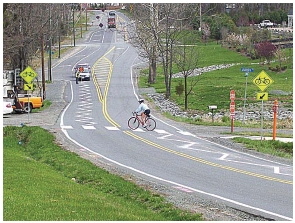 Photo depicting zig zag markings in the middle of each lane of a two-lane roadway on the approach to a mixed use (pedestrian and bicycle) crossing in a parklike suburban area.