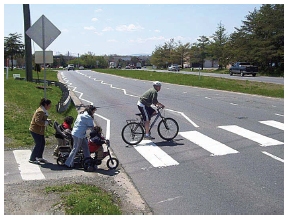 Photo depicting zig zagging edge line and lane separation line markings on the approach to a crosswalk that traverses a four-lane highway separated by a median. A bicyclist and several women with children on small bikes or in carriages are entering the crosswalk.