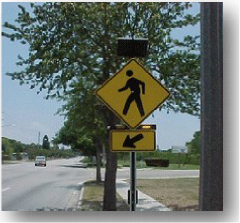 Figure 1. Photo. RRFB with two forward-facing LED flashers and a side-mounted LED flasher. A pole mounted at the side of a roadway near a crosswalk bears the W11-2 pedestrian warning sign, which shows a silhouette of a person walking to the left. The sign is a yellow diamond with a black border, and the silhouette is black. Below the sign, there is a rectangular rapid-flashing beacon (RRFB) with two yellow lights, and the light on the right is activated. Below the sign, there is a rectangular yellow sign with a black border and a black arrow pointing diagonally downward to the left toward the crosswalk. Above the W11-2 sign, there is a photovoltaic panel.