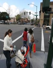 A woman waits to cross the street at a crosswalk with a baby in a stroller and two children.