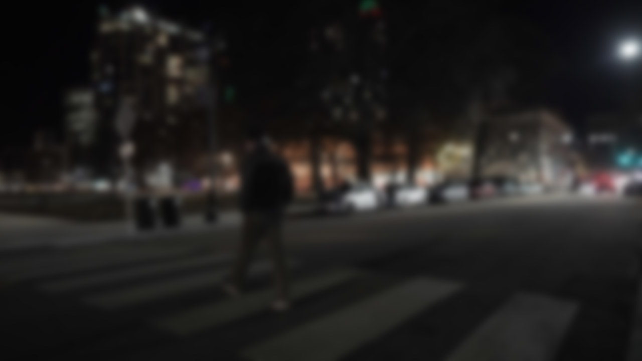 Blurry photo of man walking across in a midblock crosswalk at night in an urban setting. Lighting is dim.