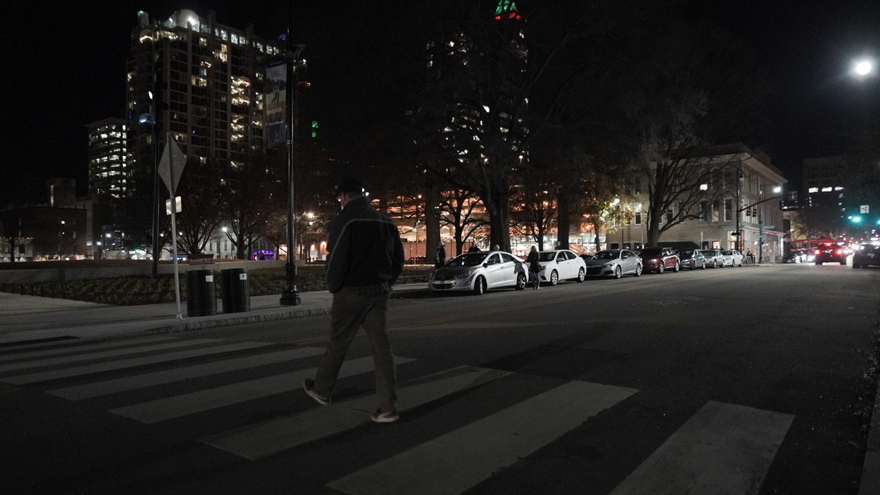 Man walks across a mid-block crosswalk in an urban setting at night. The lighting is low and it is hard to see the pedestrian.