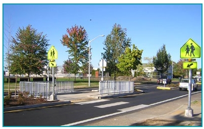 Photo of a raised median with a pedestrian refuge protected by white metal fencing that is about 3 feet in height.  The pedestrian channel is in the shape of a 2, reflecting an offset crosswalk design.