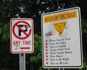 Photo.  A no parking sign and a trail etiquette sign.  The trail etiquette sign is confusing and the small text size is not readable at bicycle travel speeds. Furthermore, the no parking sign discourages cyclists from stopping.