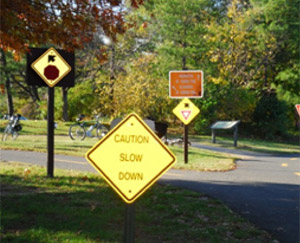 Photo.  The intersection of two shared use paths.  There are seven signs shown on one approach presenting users with multiple messages, which may be confusing or distracting.