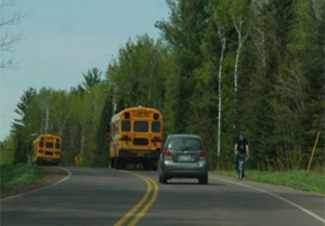 Photo.  A cyclist riding against traffic, on the shoulder of a rural two-lane road that only has a shoulder on one side.