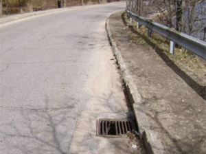 Photo.  A rural road with a grate on the right edge of the roadway.  The grate pictured at has both longitudinal openings, which could catch cyclists tires, and is damaged, leaving even wider openings.  