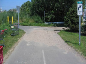 Photo.  A shared use path at the intersection with another shared use path.  Gravel and debris that has been carried by storm water and deposited at the location due to poor drainage covers the entire path.