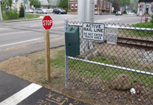Photo.  A bicycle facility runs parallel to train tracks, both of which intersect a two lane roadway.  There is a chain link fence immediately to the right of the bicycle facility that separates the facility from the tracks.  A stop sign is located along the bicycle facility, at the intersection with the roadway, but is not installed at the appropriate offset or height from the pavement edge, increasing the potential of a sign strike. Visibility of the sign is also obstructed by the fence and box. Placement of the fence adjacent to the path also restricts usable space.
