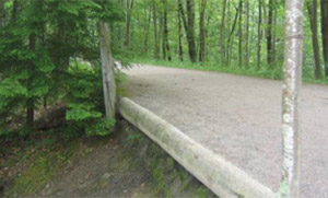 Photo.  A gravel shared use path located in the woods, with an unprotected, sharp drop-off that is non-traversable by cyclists.