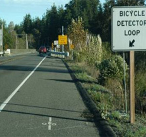 Photo.  A two lane road with a bicycle shoulder that ends at a bridge.  There is a bicycle warning system, that includes a pavement marking and a sign indicating the location of the bicycle detector loops that is used to alert motorists of the presence of cyclists on a bridge as the bikeable shoulders do not continue over the bridge.