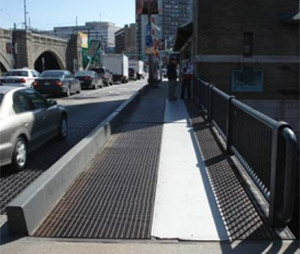 Photo.  A bridge with motorized vehicles to the left, a short physical barrier to the right, with a path on the far right for non-motorized use.  Approximately two-thirds of the path is a metal grate while the far right third is a metal plate.  The transition to the metal plate is jagged, possibly from snow removal, rather than having a smooth transition.  