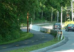 Photo.  A shared use path to the left of a two lane rural road that is separated by a vegetated strip and, in a portion, by guardrails and a concrete barrier.  The guardrail has a sharp edge that may pose an injury risk in the event of a fall.