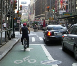Photo.  One leg of an urban signalized intersection that is comprised of a multilane one-way roadway with a bike lane in the far-left lane.  The photograph shows a cyclist waiting for a dedicated bicycle traffic signal phase.