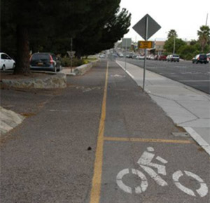 Photo.  A two-lane side path with a sidewalk to the right and the roadway to the right.  There are yellow solid lines at driveways and cross streets.  These pavement markings are to illustrate the intent for cyclists to stop at driveways and cross streets (the markings do not conform to the MUTCD).