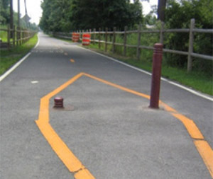 Photo.  Two brown bollards in the center of a shared use path.  The bollards are moveable and the bollard on the left has been retracted to just above the pavement surface. This may present visibility issues for cyclists using this facility as retraction reduces the bollards conspicuity.  Also, appropriate bollard positioning typically involves installing either one or three bollards positioned along the centerline and edge lines to minimize lane positioning issues for bicyclists that may lead to head-on bicycle crashes. For example, the use of two bollards may cause a situation where two head-on cyclists may choose to use the center gap at the same time and lead to a head-on collision.