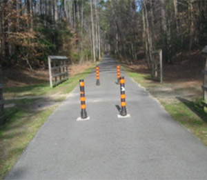 Photo.  Two sets of two black bollards with orange striping in the center of a shared use path.  The separation between the bollards allows for motor vehicle access.  Appropriate bollard positioning typically involves installing either one or three bollards positioned along the centerline and edge lines to minimize lane positioning issues for bicyclists that may lead to head-on bicycle crashes. For example, the use of two bollards may cause a situation where two head-on cyclists may choose to use the center gap at the same time and lead to a head-on collision.