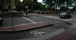 Photo.  A bicycle lane along a two lane roadway transitioning to a shared lane on a perpendicular street.  At the transition point the bicycle lane is separated to the right of the roadway by a brick island.  The transition lane includes bicycle pavement markings and has room for bicyclists to wait until the signal changes so they can make a left onto the shared lane.  The crossing is signalized to assign the right-of-way between motorists and cyclists. Pedestrians and cyclists are also provided separated crossing paths.