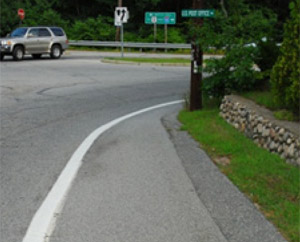 Photo.  A wide, bikeable shoulder on the right side of a rural two lane road.  The shoulder terminates at an unsignalized intersection that also located along a horizontal curve that has limited sight distance due to vegetation.  