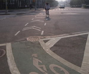 Photo.  A bike lane on the left side of a roadway that continues straight through a major intersection and transitions to a protected bikeway, also on the left side of the roadway. Chevrons on the pavement help guide cyclists and show motorists the path provided for cyclists through the intersection (note that the chevron pavement markings do not conform to the MUTCD).