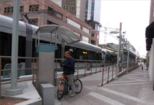 Photo.  A cyclist is using an automated fare collector at the access ramp for a transit stop and is partially blocking access to the platform.  Furthermore, the width of the access ramp to the platform is narrow and may not adequately accommodate peak demands, especially when pedestrians with mobility restrictions and cyclists are both present.