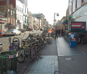 Photo.  An urban sidewalk that has been widened to allow space for bike racks in the street furniture zone. The added space allows for adequate space for bicycle storage along with space to access a busy subway station.