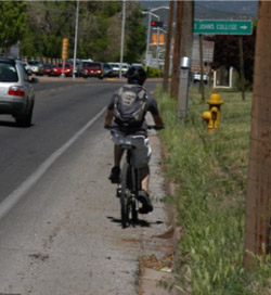 Photo.  The photo shows a person riding their bicycle along the shoulder of a busy highway.  The shoulder is covered in gravel and the cyclist is riding in the same direction of traffic.