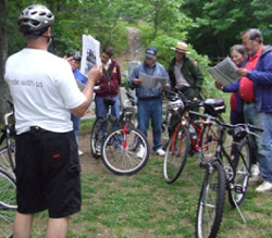 Photo.  A team prepares for an RSA on a shared use path.  The individual leading the team is standing in front and facing the rest of the team who are standing next to their bikes and holding materials relevant to the RSA.