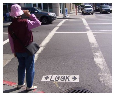 Photo of a pedestrian standing on a curb at a crosswalk where the word 'LOOK' had been stenciled onto the pavement at the beginning of the crosswalk. The Os in the word LOOK have been painted to look like two eyes.