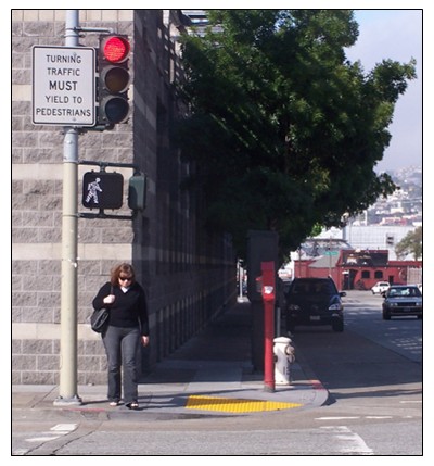 Photo of a pedestrian signal at a crosswalk showing the 'Walk' phase while the traffic signal remains in the red phase. A sign indicates traffic must yield to pedestrians in the crosswalk.