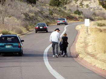 Couple with infant in stroller walk on shoulder along a one-lane road