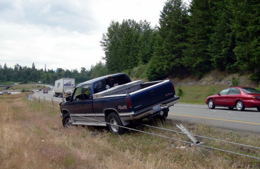 Photo: This photograph, taken from the median of a four-lane divided highway, shows a pickup truck restrained by a median cable barrier. The median cable barrier consists of two horizontal metal cables supported by metal posts. The pickup truck pointing towards opposing traffic at an acute angle. One of the metal cables that comprise the median cable barrier is located underneath the truck, while the other is running along the driver side of the vehicle.