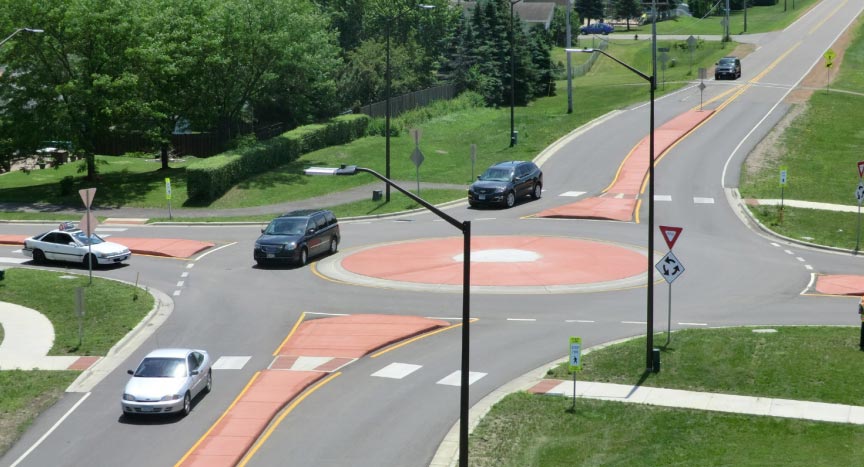 Photo: This photograph shows a single-lane roundabout. Each of the four legs approaching the roundabout is a yield-controlled, two-lane roadway. Each approach leg features a high visibility crosswalk. Raised medians are present in the vicinity of the roundabout but not beyond the extents of the roundabout. The roundabout island appears to be traversable by trucks and other large vehicles. 