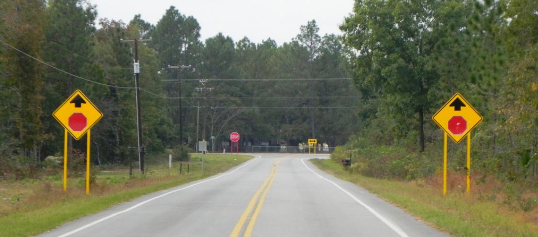 Photo: This photograph, taken along the centerline of a two-lane undivided roadway, shows countermeasures present on the minor leg of a T-intersection. On both sides of the roadway, W3-1 Stop Ahead warning signs are present. Each W3-1 sign is supported by two metal posts covered in yellow reflective material.