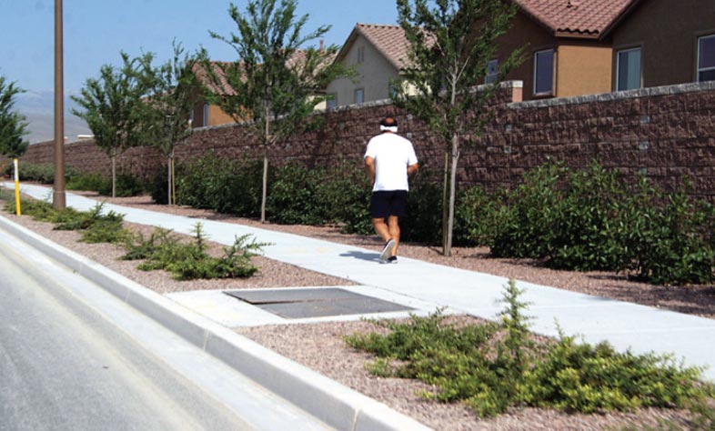 Photo: This photograph, taken from the roadway, shows a sidewalk in a residential area. The concrete sidewalk is separated from the roadway by a concrete curb and landscaping area. A pedestrian can be seen running on the sidewalk.
