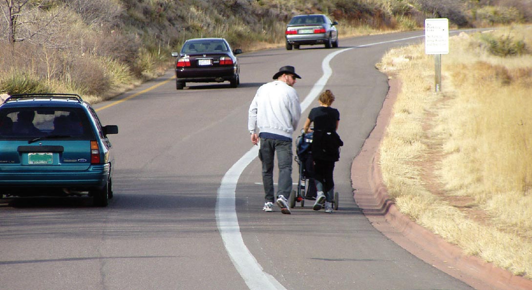 Photo: This photograph, taken from the roadway, shows a paved shoulder being used as a walkway by two people in a rural area. Vehicles on the roadway are shown traveling in the same direction, indicating that the roadway is a one way or a divided facility. The paved shoulder and general travel lane are separated by a wide, solid white line. The outside of the paved shoulder is bordered by a concrete curb. Beyond the curb, a footpath is visible in the adjacent grass. 