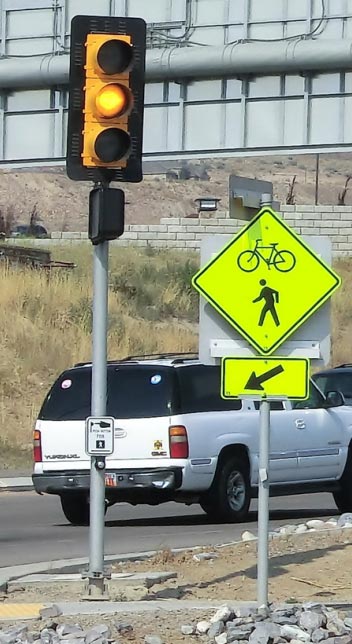 Photo: This photograph shows a traffic signal showing YELLOW at a trail crossing. The traffic signal is mounted with a black backplate onto a pole. Next to the pole supporting the traffic signal is a bicycle and pedestrian shared warning sign with supplemental directional arrow plaque. A white SUV is visible driving along the roadway in the background of the photograph.