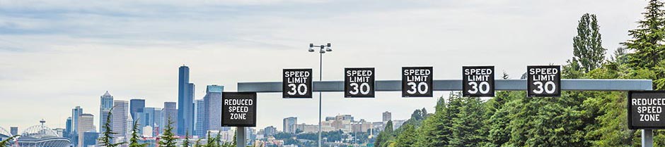 Variable speed limit signs are positioned on a crossbar over several lanes of a highway. The city of Seattle, WA, is in the distance.