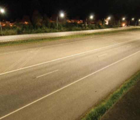 Photo: : This nighttime photograph shows a series of seven streetlights illuminating a section of a multilane highway.
