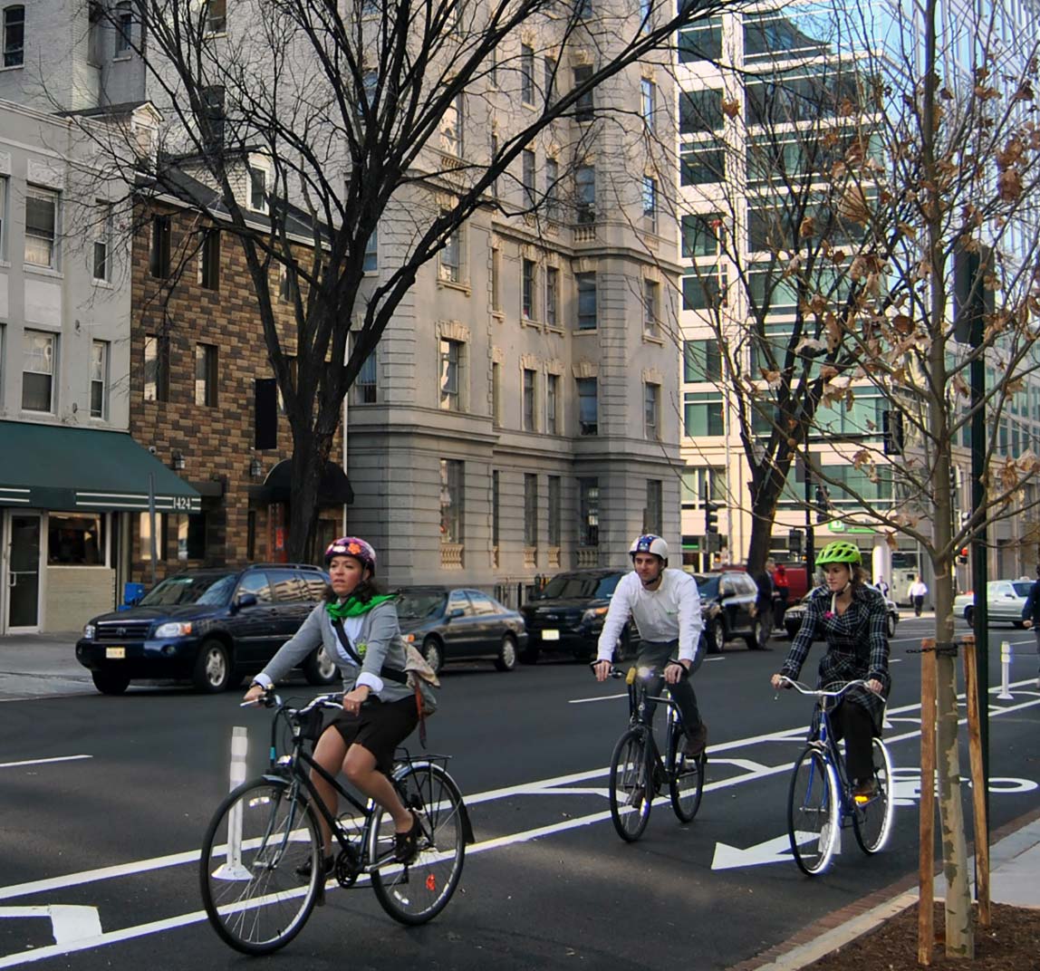 Photo: This photograph, taken from the edge of the roadway, shows three bicyclists traveling in a buffered bicycle lane toward the foreground of the image in an urban setting. To the left of the bicycle lane are two general travel lanes with on-street parking. The bicycle lane is separated from the roadway with white pavement markings and vertical posts. The bicycle lane is marked with a helmeted bicyclist symbol and directional arrow. To the right of the bicycle lane is concrete curb and sidewalk.