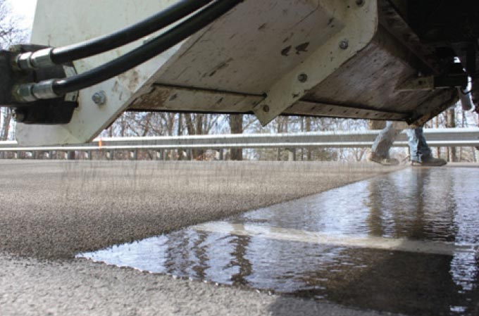 Photo: This photograph, taken along the roadway surface, shows the application of high friction surface treatment on a roadway. A white metal ramp, which is attached to a vehicle, extends across the travel lane, spraying material onto the surface below. Underneath the metal ramp liquid is visible on the roadway surface.