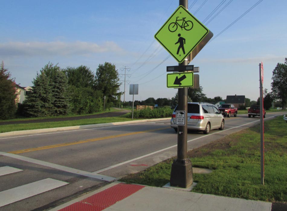 Photo: This photograph shows use of a rectangular rapid flashing beacon at a trail crossing. The crossing features a high visibility crosswalk and tactile curb ramp in the foreground of the image. The center of the image focuses on a W11-15 combined bicycle/pedestrian warning sign with rectangular rapid flashing beacon and W16-7P directional arrow plaque underneath. Below the warning sign assembly is a crosswalk button with instructional plaque.