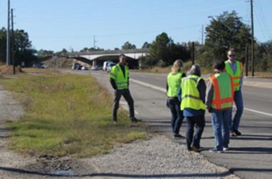 This photograph, taken from the side of the roadway, shows a group of five individuals inspecting the roadway. The five individuals are standing in the paved shoulder of the roadway and wearing reflective safety vests.