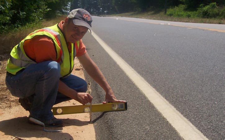 Photo: This photograph, taken along the edge of a roadway, shows the angle of SafetyEdge relative to the roadway surface. The photograph shows a person holding a level perpendicular to the edge of the roadway. There is no backfill material adjacent to the paved surface, revealing the SafetyEdge below. The SafetyEdge consists of a slope of asphalt installed at approximately a 30-degree angle to the horizontal.