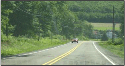 Photograph of a two-lane roadway in a rural area. A car is coming from a horizontal curve. A 4-inch edge line is installed on both sides of the roadway prior to the curved section.