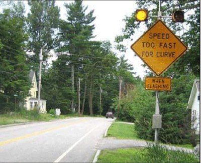 Photograph of a flashing beacon on a warning sign, which is installed prior to a curve of a two-lane roadway in a residential area. The texts on the warning sign are read as Speed too fast for curve when flashing.