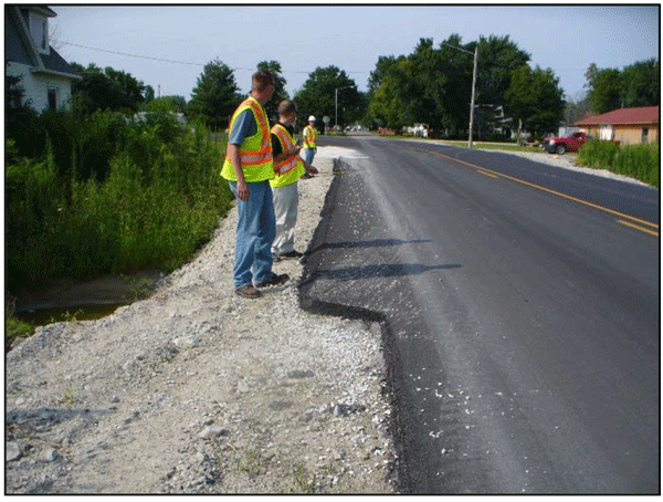 Photograph showing a shoulder paving operation in the field. The operation is conducted on a two-lane roadway in a suburban area. Three workers wearing safety vests are standing off the road and supervising the operation.