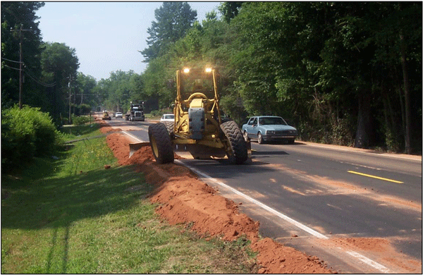 Photograph of a motor grader backfilling against newly installed Safety EdgeSM on a two-lane roadway. The roadside vegetation is brought up even with the pavement surface.