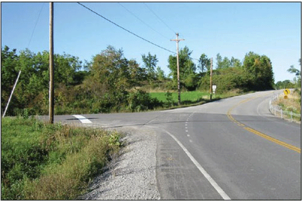 Photograph of a three-leg intersection in a rural area. The three-leg intersection is located within a curved section of a two-lane roadway. The roadway has edge lines and centerlines. Dotted edge line extensions are installed on the cross road.