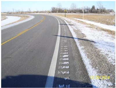 Close-up photograph of a narrow paved shoulder (2 feet). It is a two-lane roadway in a rural area. On the shoulder of the curve, rumble strips are installed adjacent to the edgeline and safety edge is installed along the edge of the roadway surface.